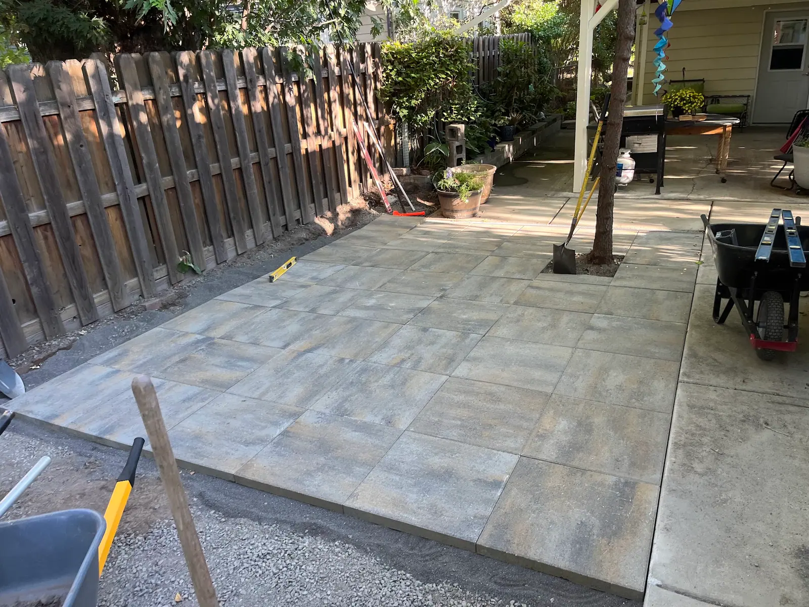 Paver patio in a fenced backyard with tools and a wheelbarrow